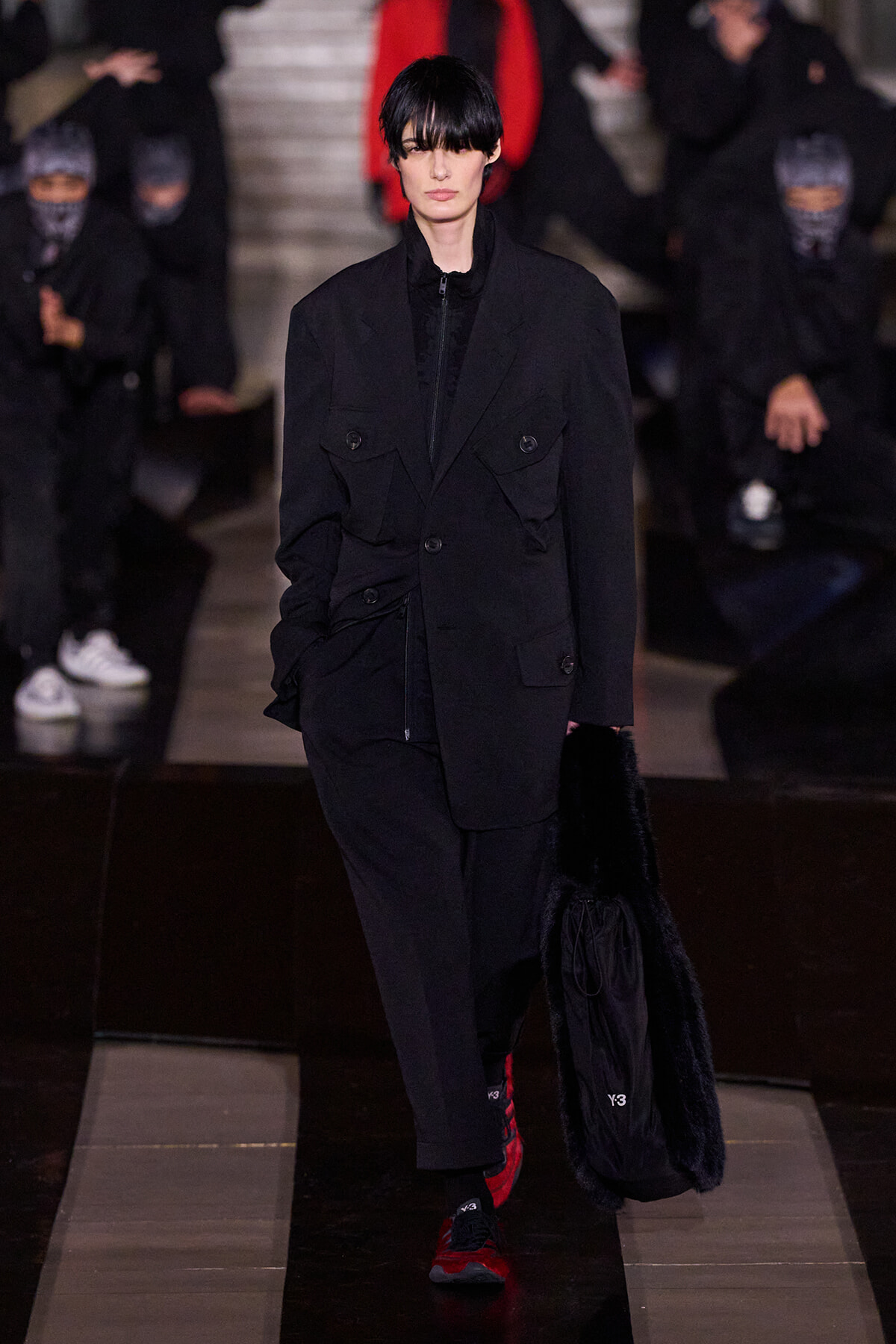 Model walks a runway in an oversized black utility jacket with multiple pockets, black pants, red sneakers, and a black fur-lined bag; dark hair clipped at the bangs with a focused expression.