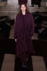 Model walking a fashion runway in a loose burgundy coat and matching dress, hands in pockets.