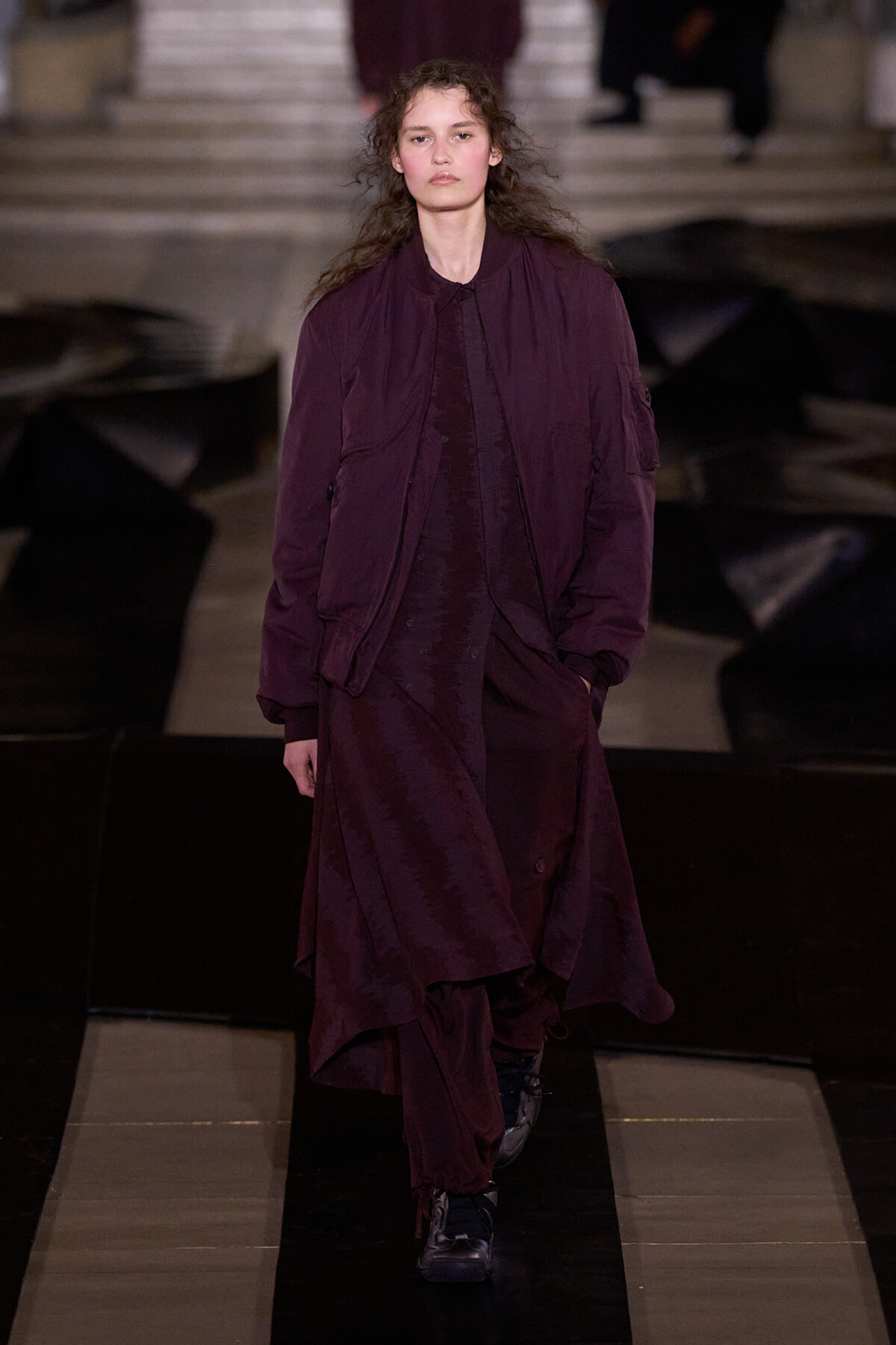 Model walking a fashion runway in a loose burgundy coat and matching dress, hands in pockets.