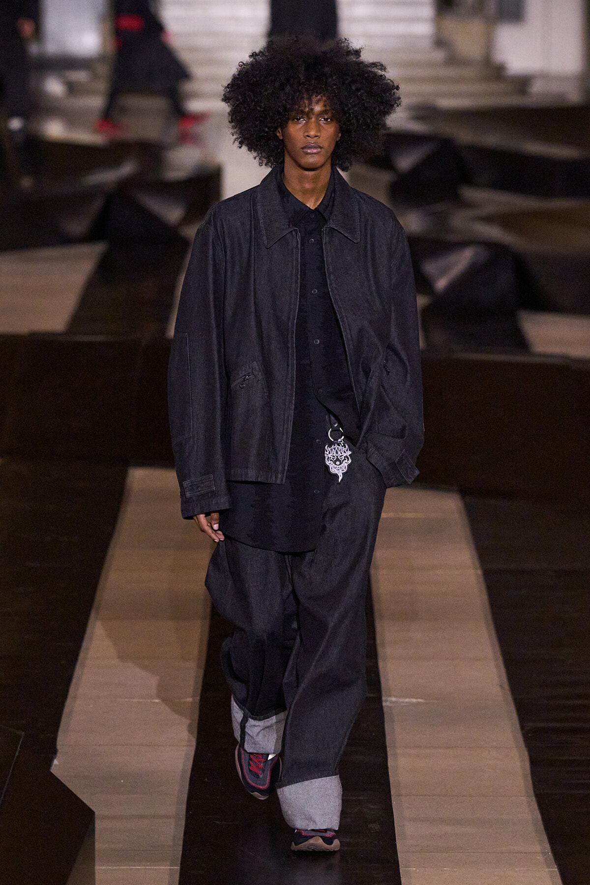 Model on a runway wearing an oversized black denim jacket and baggy jeans, with a silver pendant keychain.