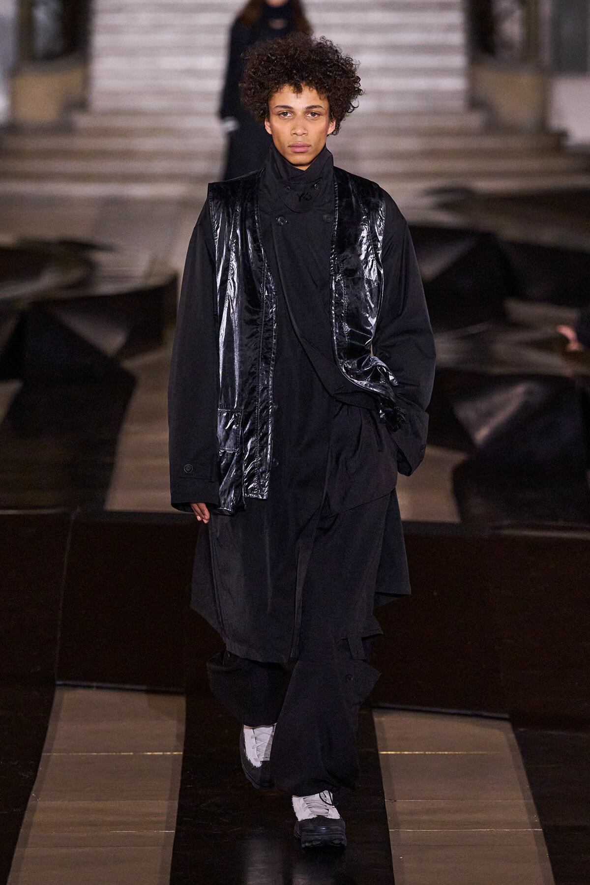 Model walking the runway in an all-black layered outfit, featuring a shiny black vest over a long black coat and white sneakers.