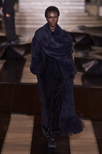 Female model walking a runway in a long navy velvet coat with an oversized textured scarf draped around her shoulders.