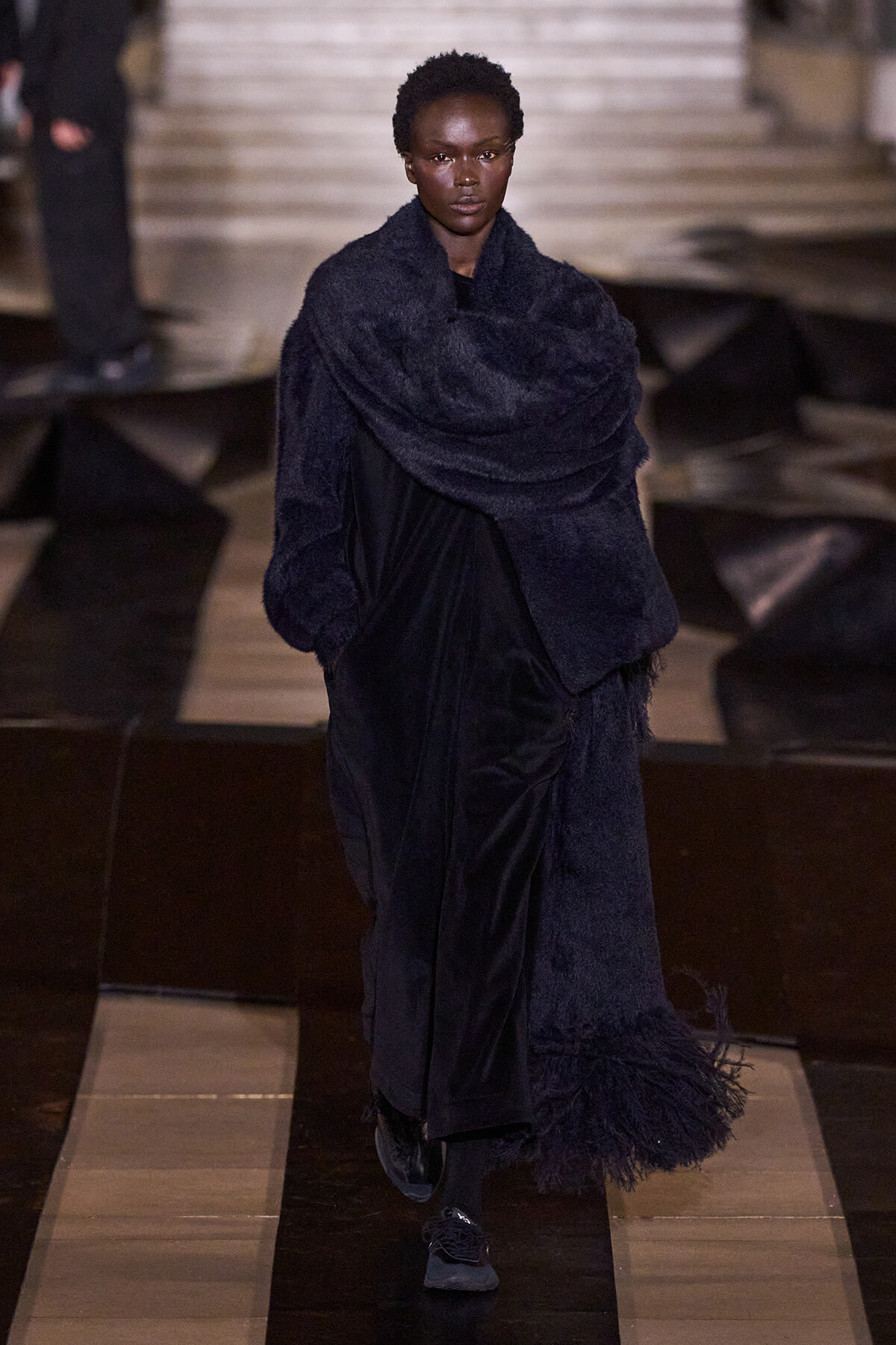 Female model walking a runway in a long navy velvet coat with an oversized textured scarf draped around her shoulders.
