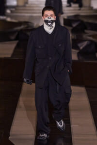 Male model walking a fashion runway in an all-black oversized jacket and cargo pants, wearing a black-and-white scarf mask.