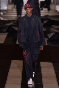 Male model wearing a dark denim jacket and pants with red graffiti accents, white sneakers, and a red-cap on a runway.