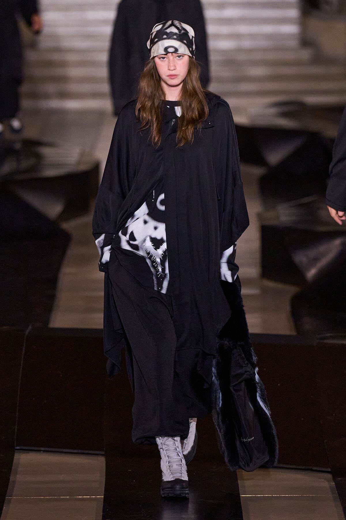 Model walking a fashion show in an oversized black cloak with a black-and-white patterned headscarf and light-colored lace-up boots on a dark runway surface in the background.