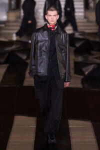 Male model wearing a black leather jacket over black clothing and a red patterned scarf walks down a fashion runway.