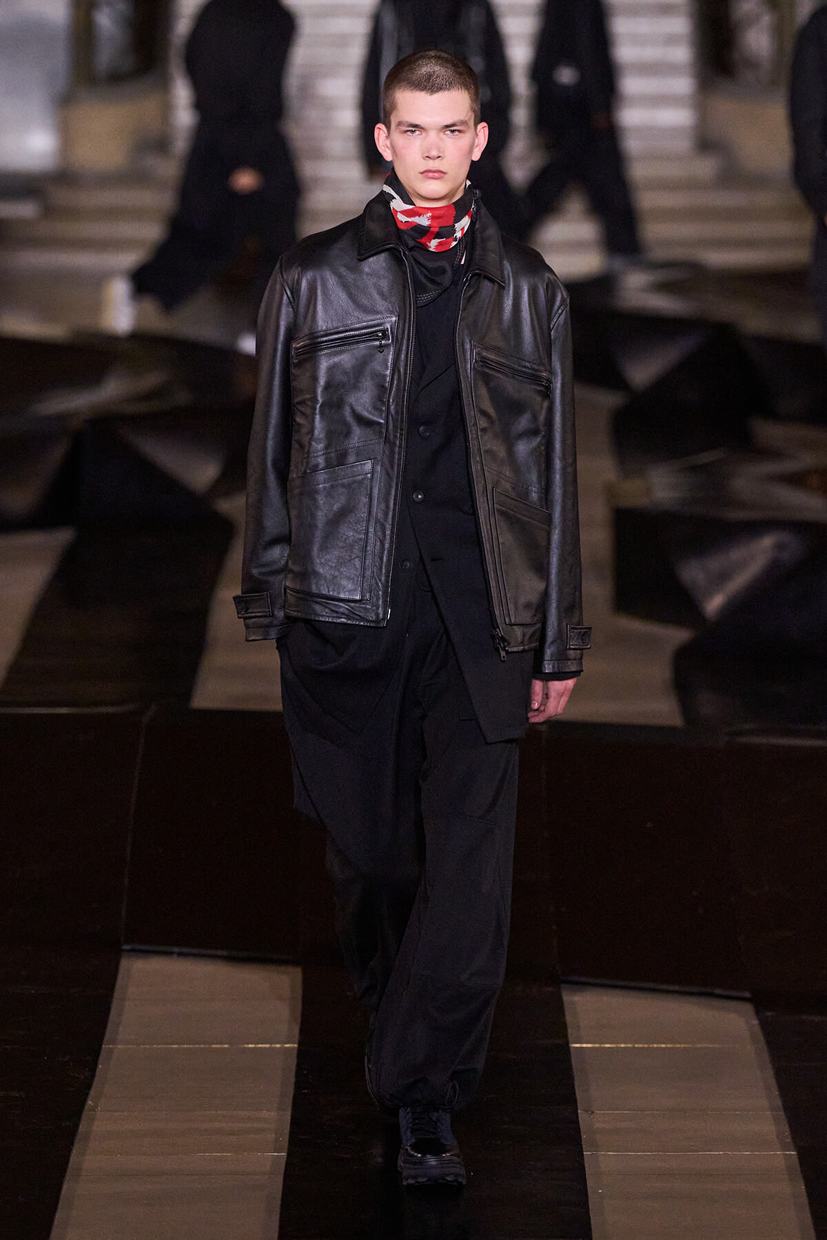 Male model wearing a black leather jacket over black clothing and a red patterned scarf walks down a fashion runway.