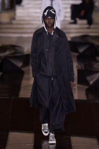 Model walking a fashion runway in an all-black, oversized coat with a hood and white‑trim sneakers.