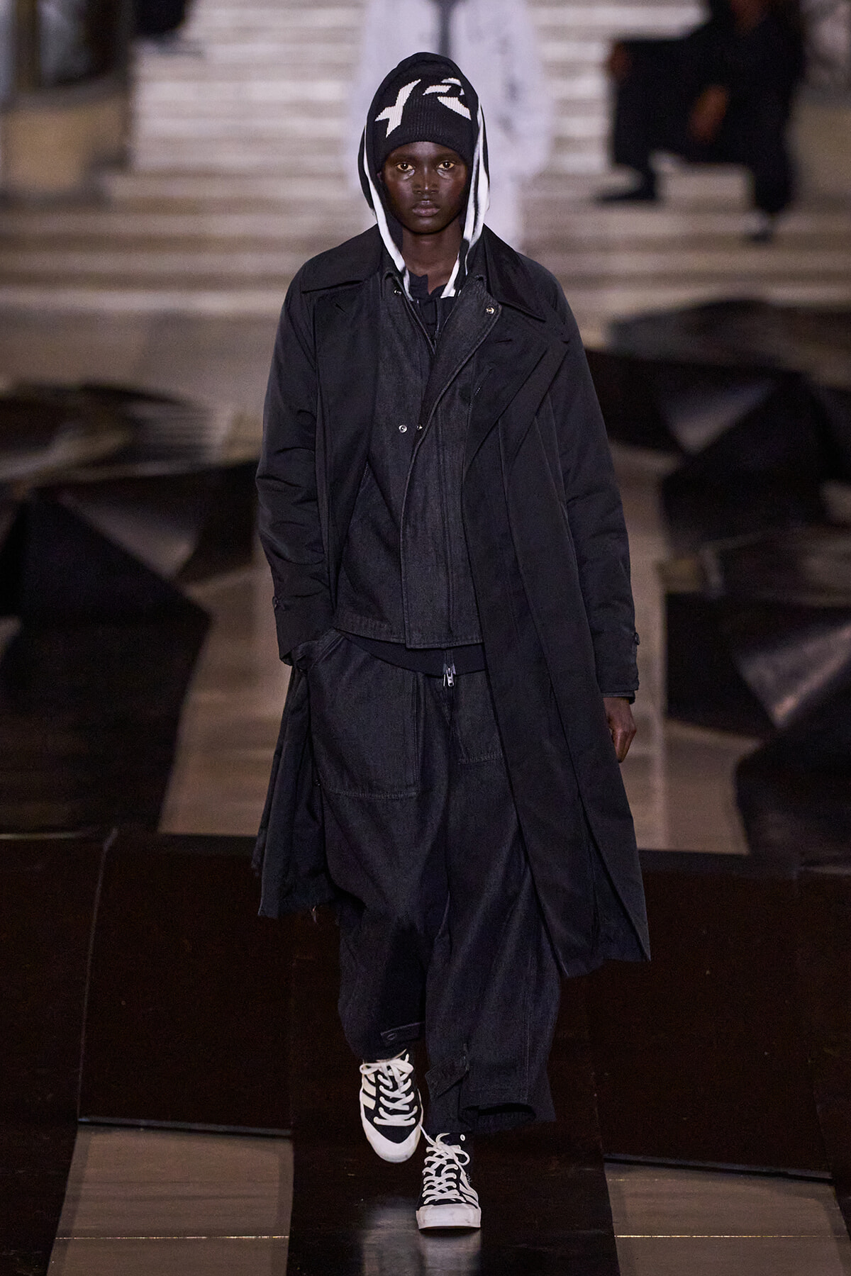 Model walking a fashion runway in an all-black, oversized coat with a hood and white‑trim sneakers.