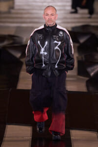 Male model walking a runway in a black leather jacket with white graffiti-like symbols and red piping, paired with dark cargo pants and red cuffed legs.