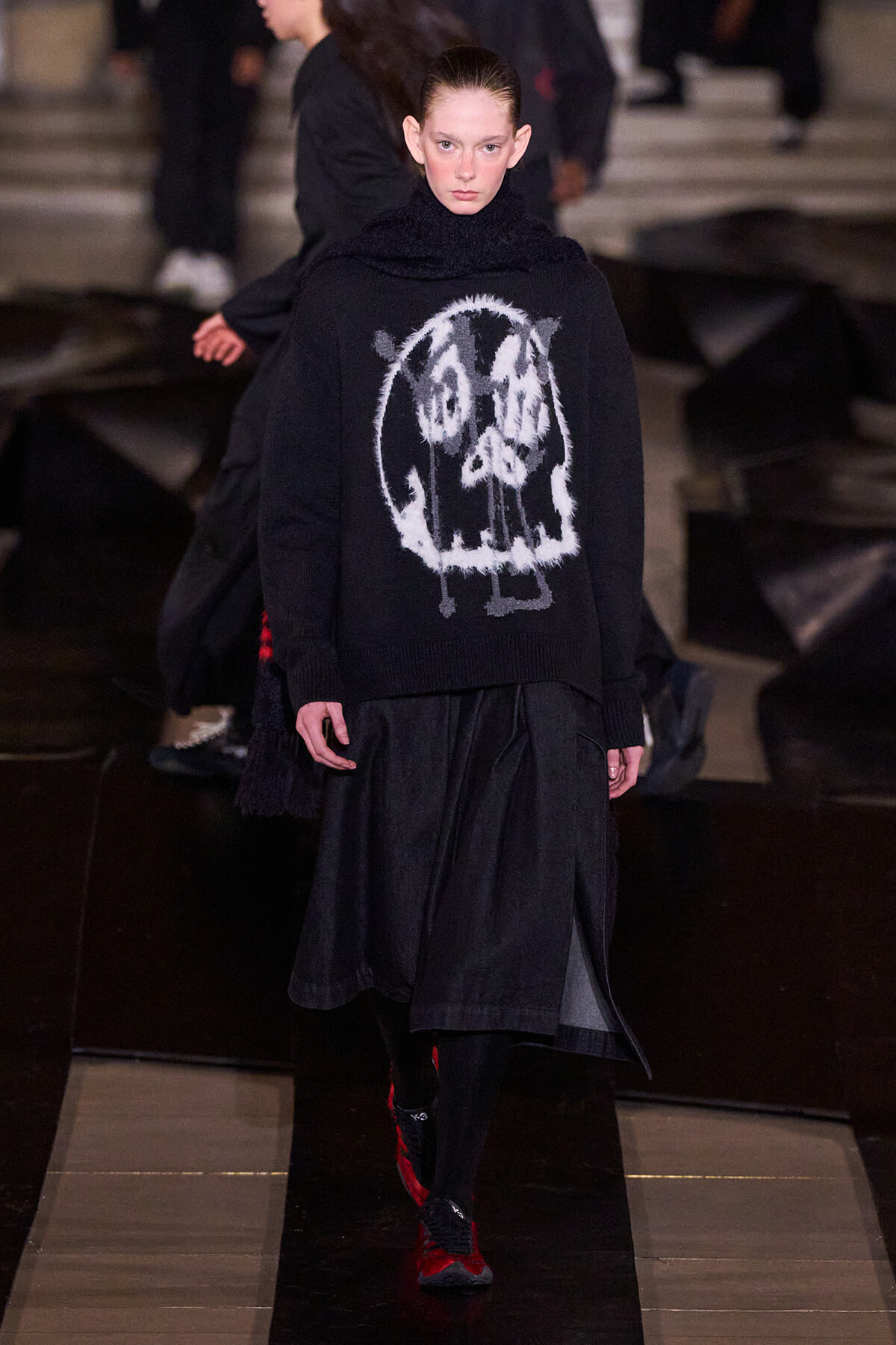 Model walking a fashion show wearing a black knit sweater with a white abstract graphic.