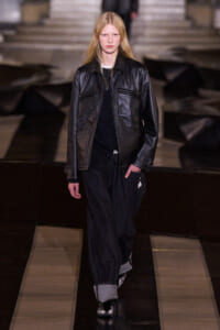 Model walks the runway in a black leather jacket over a dark outfit, with wide rolled-cuff pants, stepping forward.