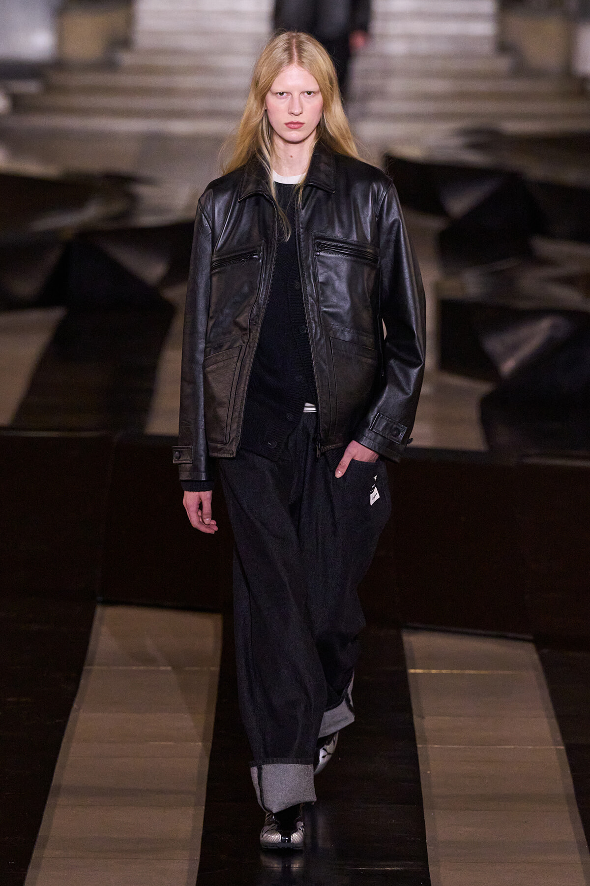 Model walks the runway in a black leather jacket over a dark outfit, with wide rolled-cuff pants, stepping forward.