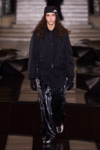 Runway model in all-black streetwear: oversized zip jacket, loose shiny pants, gloves, and a headband walking a fashion show runway.