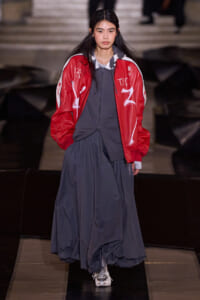 Model walking a runway in a red graphic jacket over a gray hoodie and a long navy dress with sneakers.