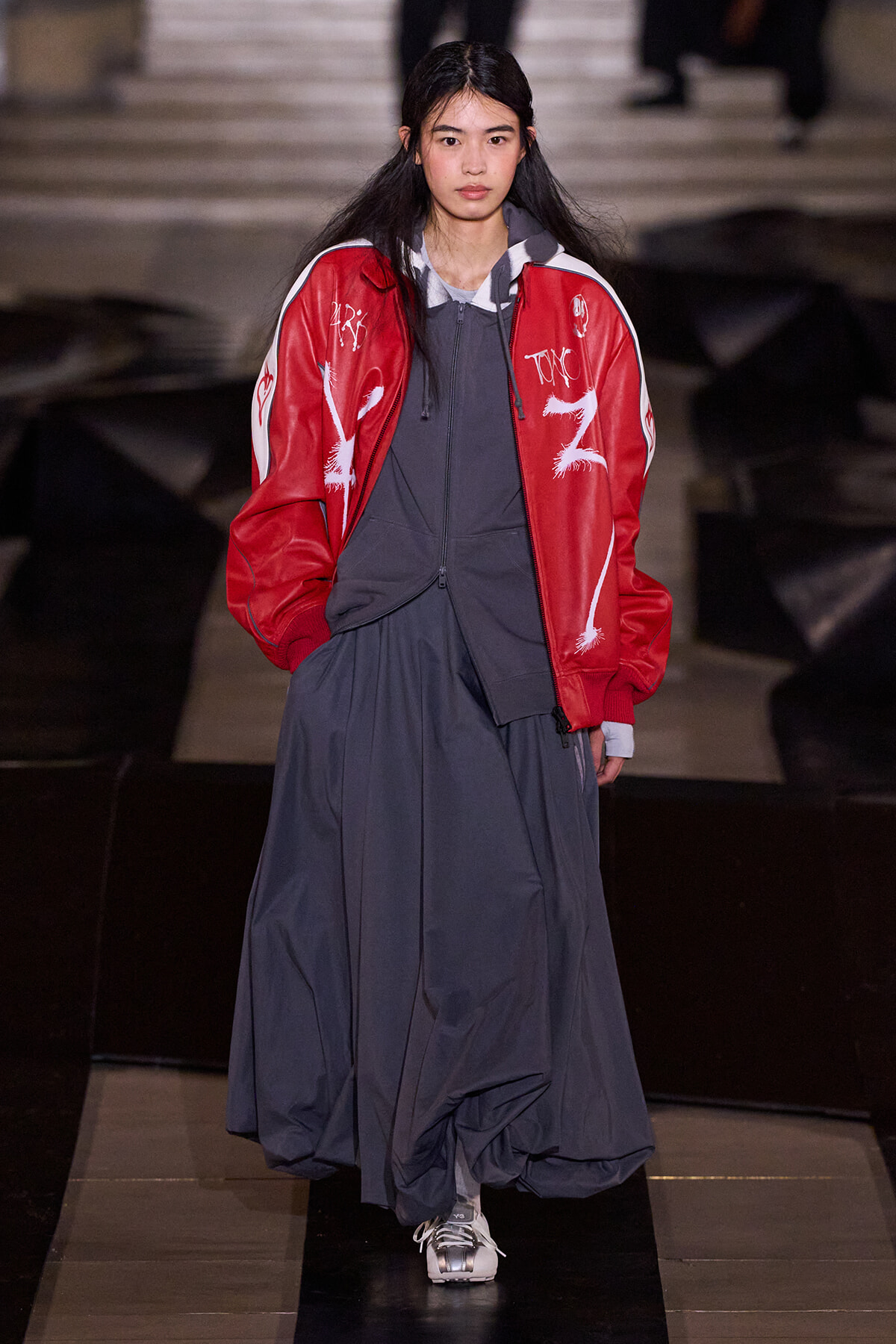 Model walking a runway in a red graphic jacket over a gray hoodie and a long navy dress with sneakers.