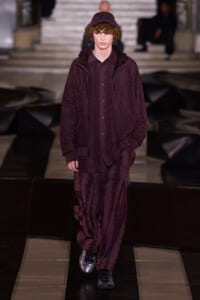 Male model walks a fashion show runway in a monochrome burgundy outfit: textured jacket, matching shirt and wide pants, wearing a burgundy cap.