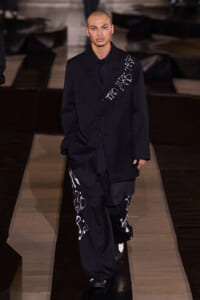 Male model walking a runway in an oversized black suit with white graffiti-style text on the blazer and pants.