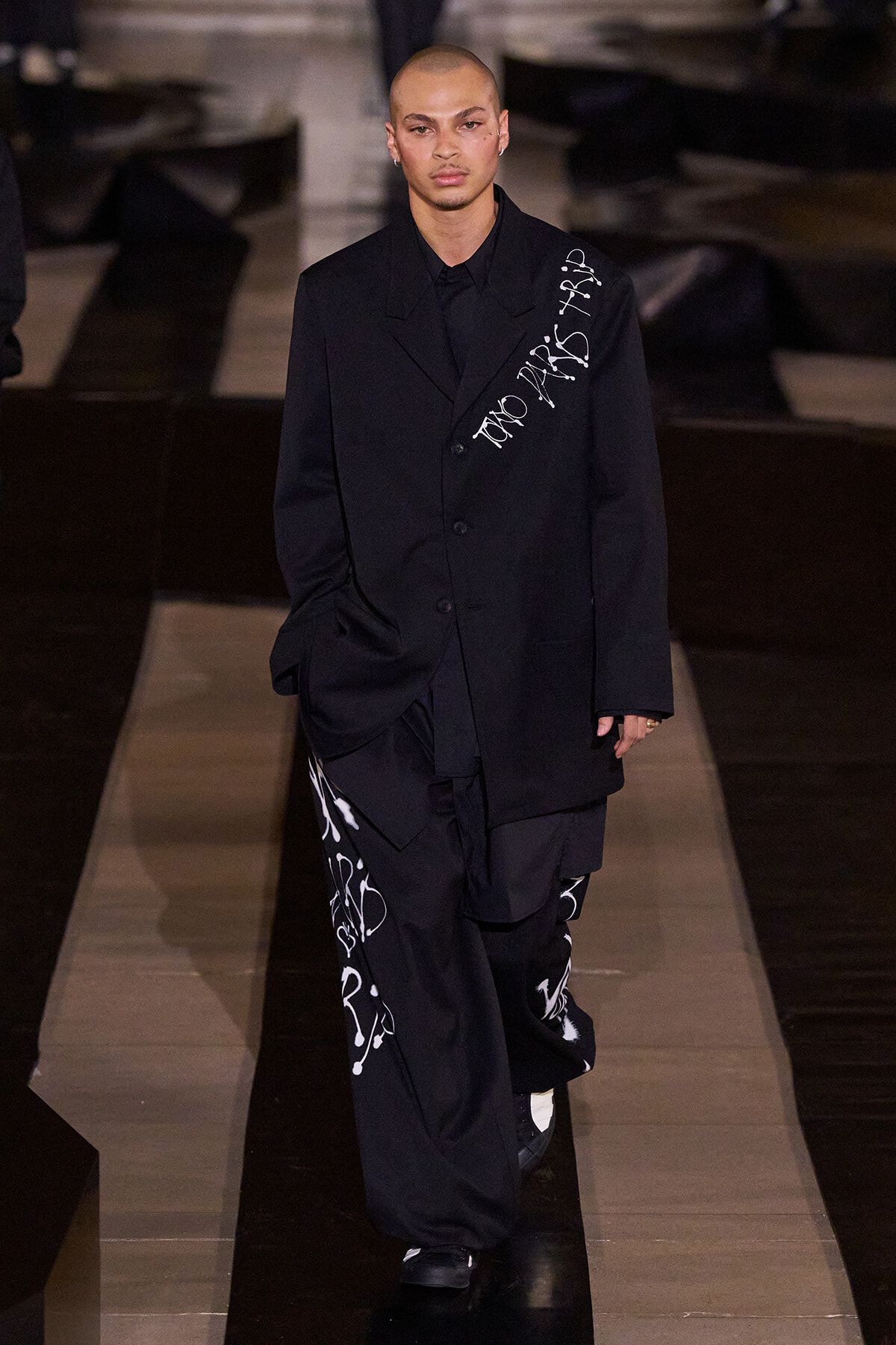 Male model walking a runway in an oversized black suit with white graffiti-style text on the blazer and pants.