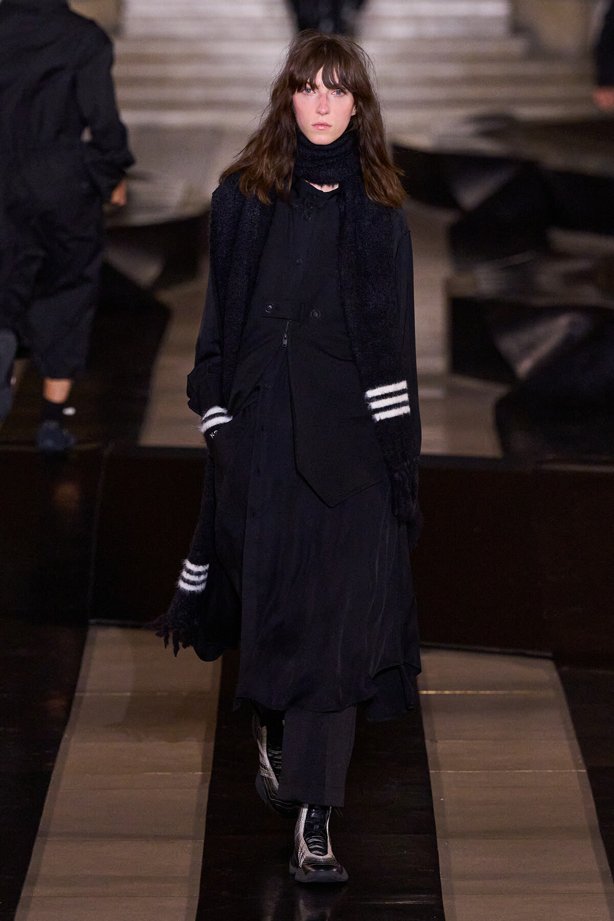 Model walking a fashion runway in an all-black, layered outfit with a long coat, scarf, and striped gloves/cuffs.