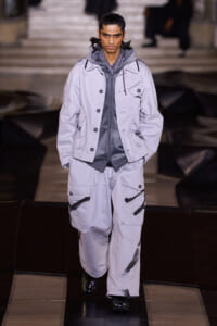 Male model walks the runway in an oversized light gray jacket and matching cargo pants with black spray-paint accents.