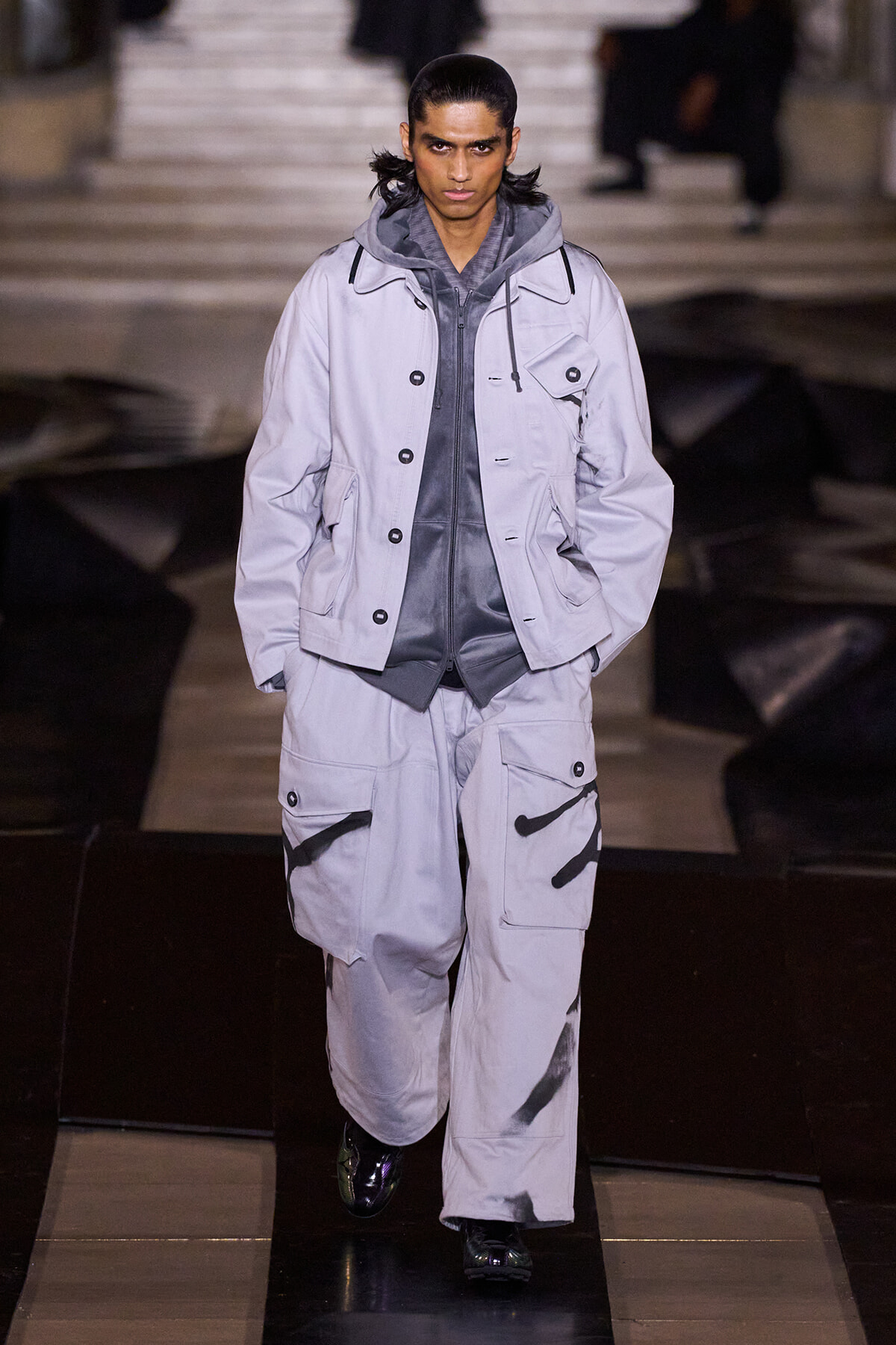 Male model walks the runway in an oversized light gray jacket and matching cargo pants with black spray-paint accents.