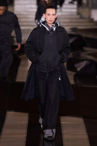 Runway model wearing an all-black, layered outfit: oversized coat, zipped hoodie, and loose denim pants with a scarf at a fashion show.