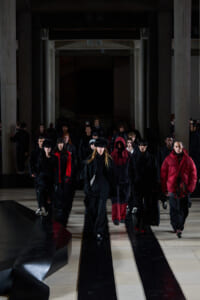 Fashion models walk down a dim runway wearing black and red outerwear inside a large hall with tall columns.