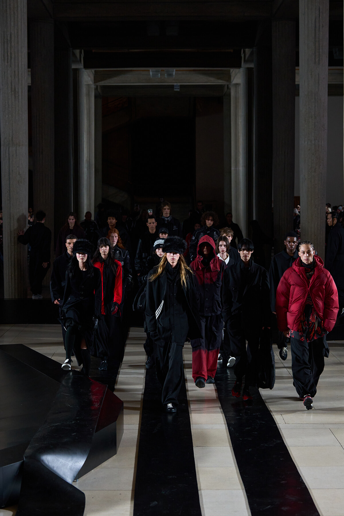 Fashion models walk down a dim runway wearing black and red outerwear inside a large hall with tall columns.
