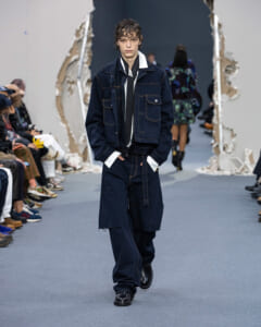 Male model walking a runway in a navy denim jacket and wide-leg jeans, white shirt cuffs visible, hands in pockets.