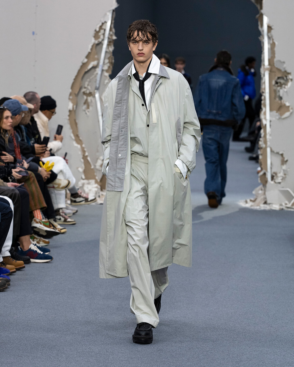 Male model walking a fashion runway in a pale gray and white layered trench coat, hands in pockets, audience along the sides.