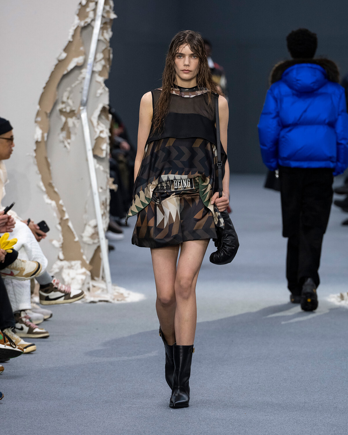 Model walking a runway in a geometric brown and black mini dress with a sheer black top, knee-high black boots, and a black shoulder bag.