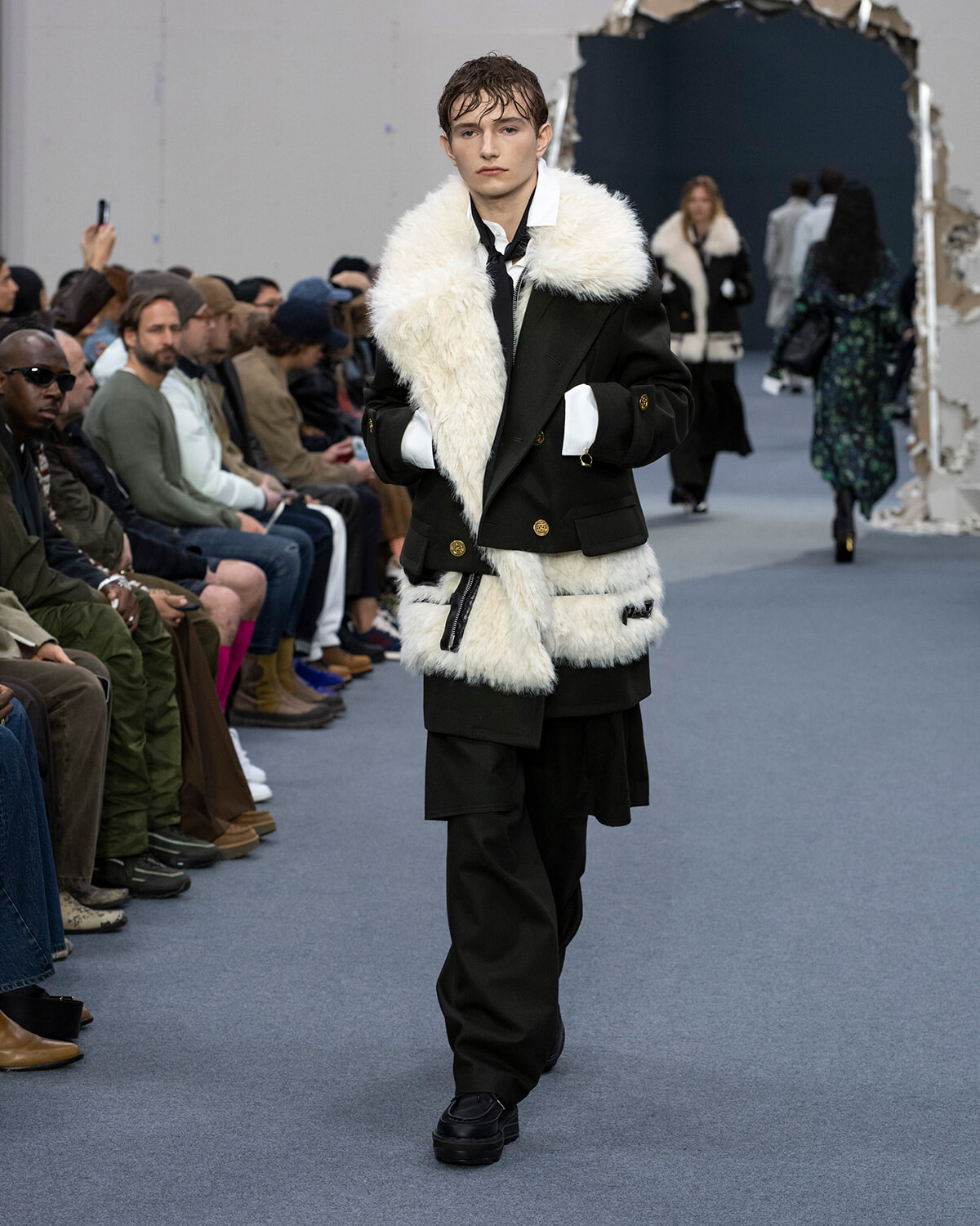 Male model walks a fashion runway in a black double-breasted coat with white faux-fur trim and panels, audience seated along the catwalk.
