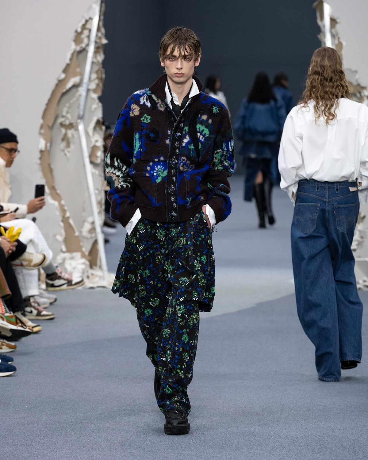 Model walks the runway in a dark floral-pattern cardigan over a white shirt and layered floral pants, stage setting with audience.
