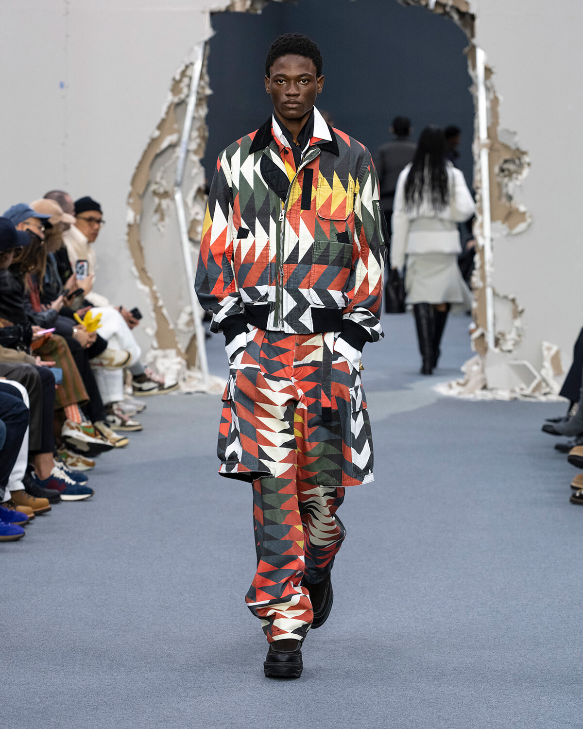 Male model on a runway wearing a matching bold geometric print jacket and pants in red, black, white, and yellow.