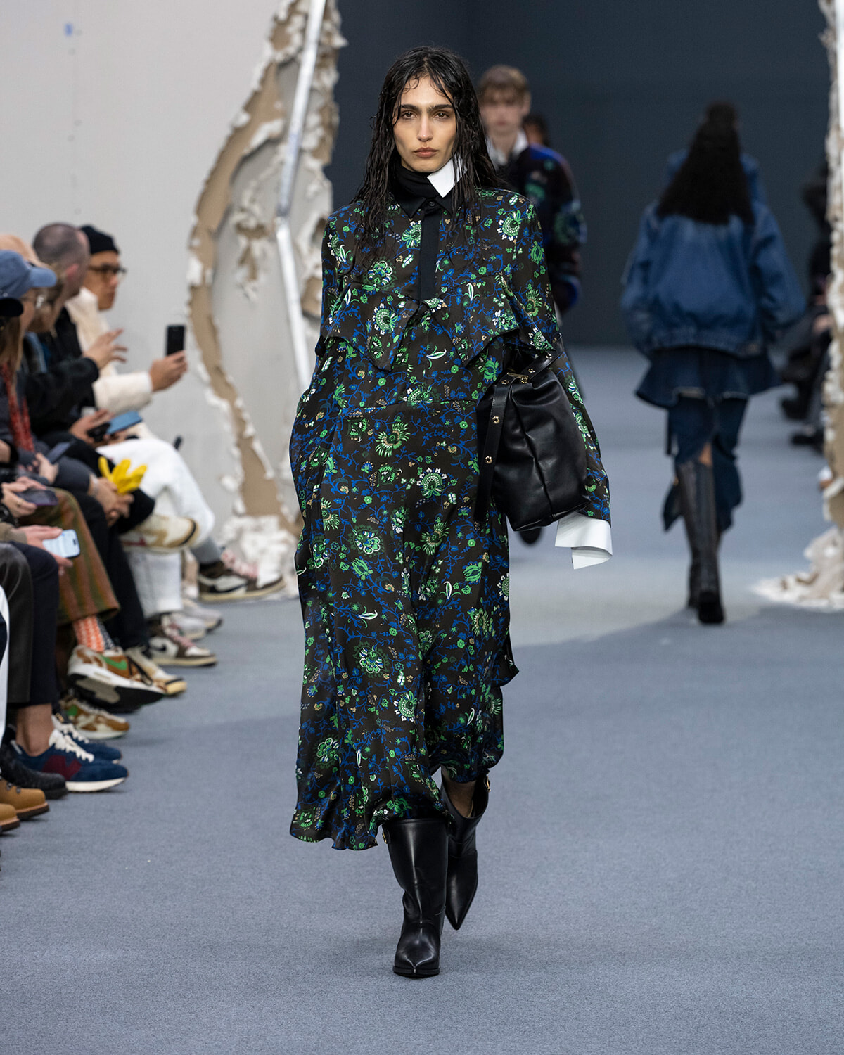 Model wearing a dark blue–green floral maxi dress with white cuffs, black boots, and a black handbag walking the runway.