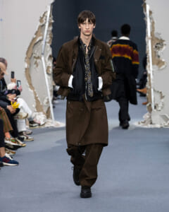 Male model walks the runway in a brown oversized coat layered over black, with a patterned scarf and matching brown pants.