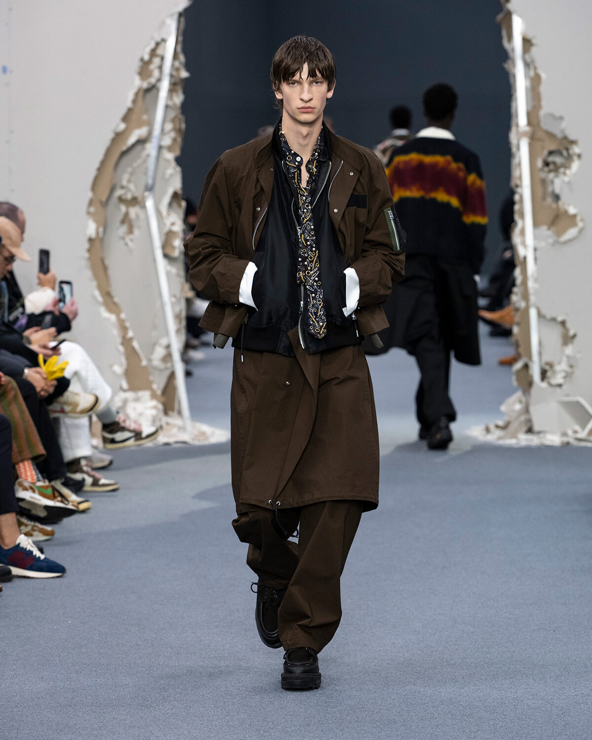 Male model walks the runway in a brown oversized coat layered over black, with a patterned scarf and matching brown pants.