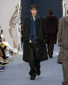 Male model walks the runway in a black long coat over a blue denim shirt and jeans, during a fashion show.
