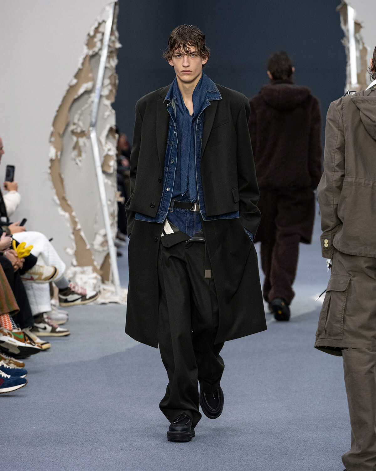 Male model walks the runway in a black long coat over a blue denim shirt and jeans, during a fashion show.