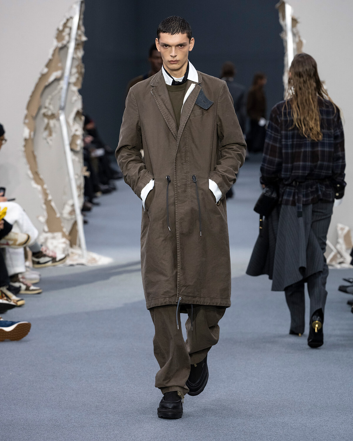 Male model walking a fashion runway in a long brown coat with drawstrings, layered over a green sweater and white-collared shirt.