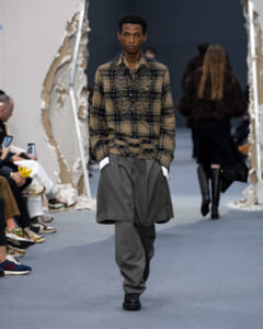 Male model walking a fashion runway in a brown plaid shirt with beaded embellishments and oversized gray layered trousers.