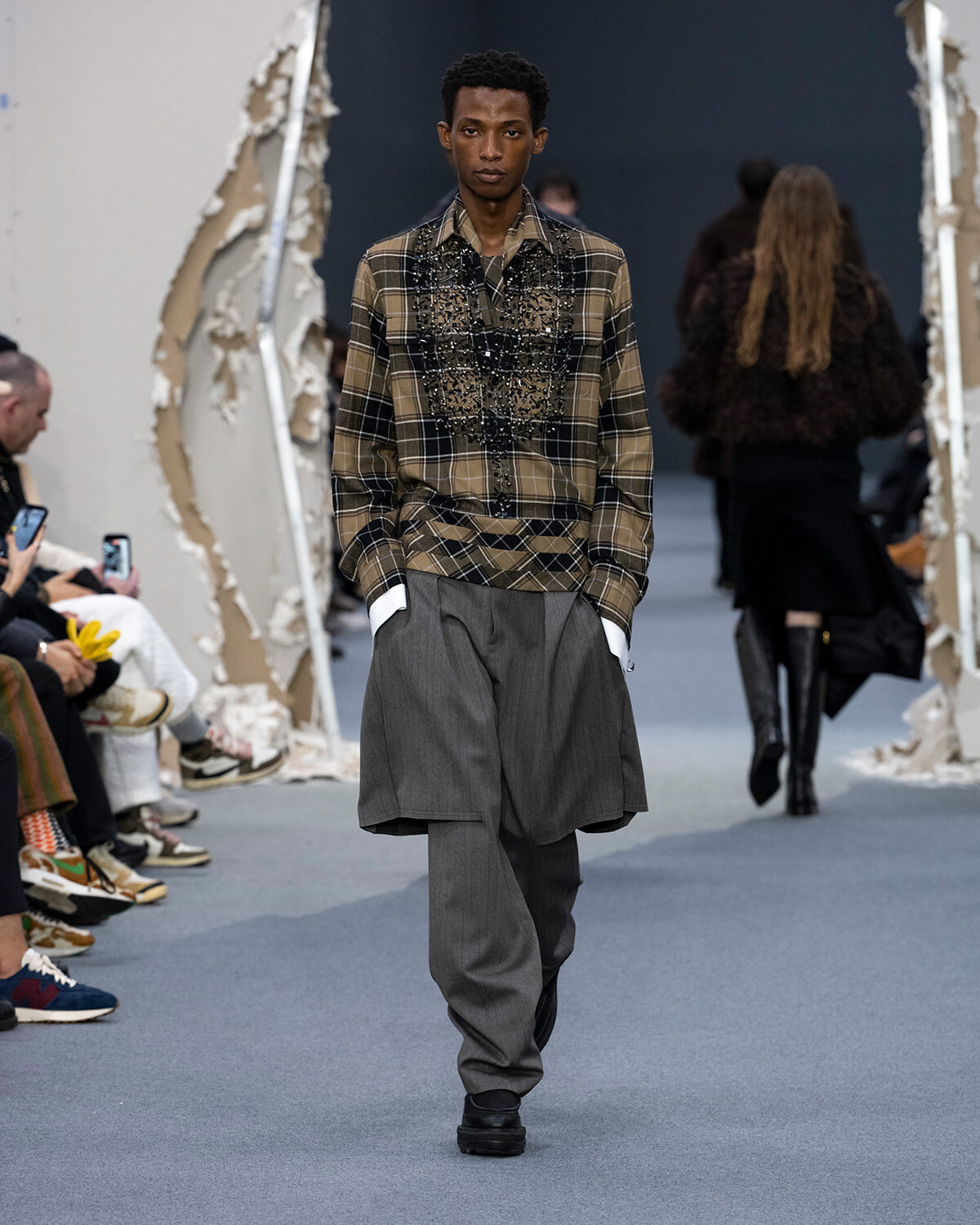 Male model walking a fashion runway in a brown plaid shirt with beaded embellishments and oversized gray layered trousers.
