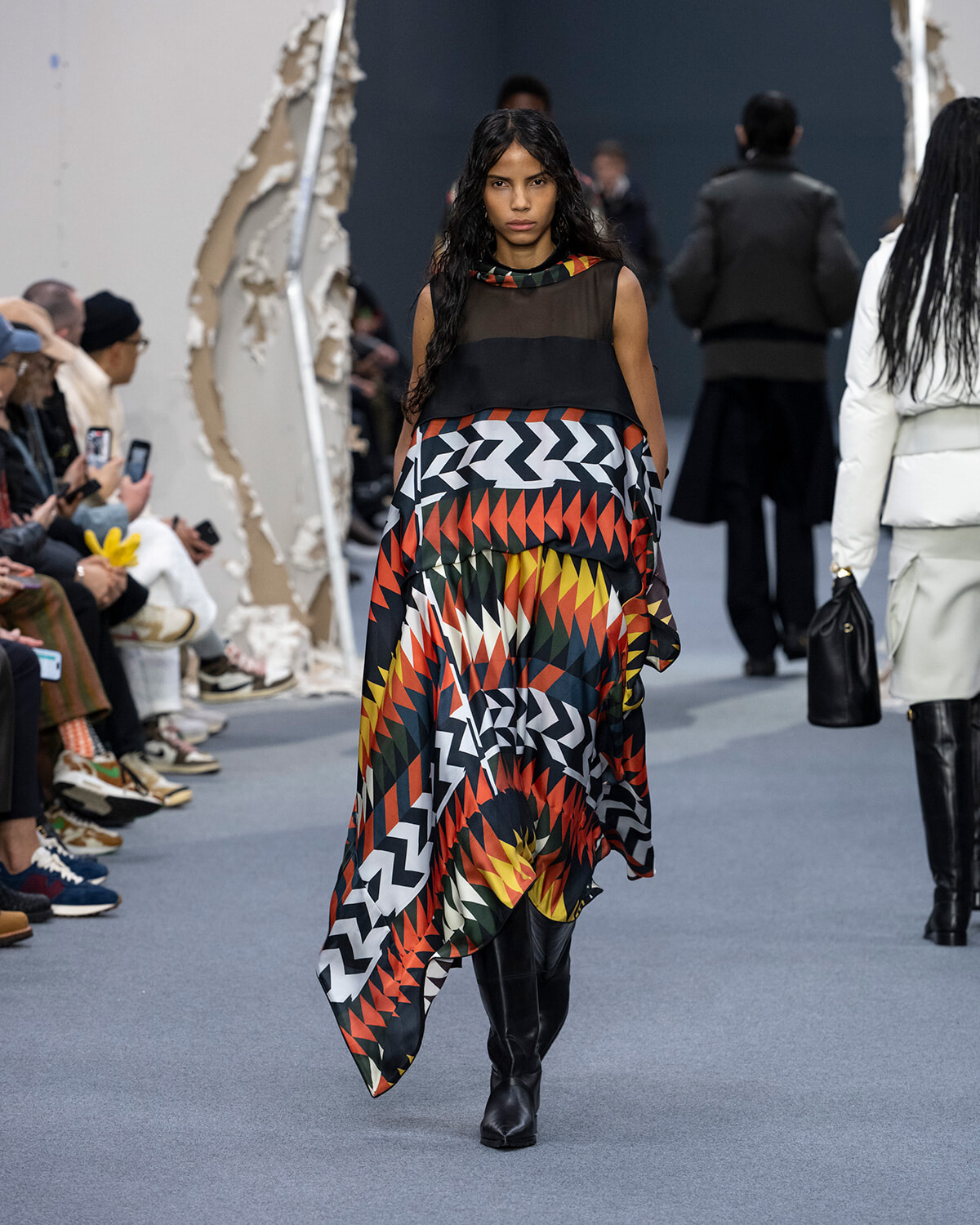 Model on a runway wearing a sleeveless sheer black top over a bold multicolored zigzag maxi dress with black knee-high boots.