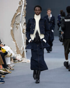 Model walks a fashion show wearing a navy denim jacket over a white turtleneck and a matching denim skirt with black knee-high boots under bright runway lights.