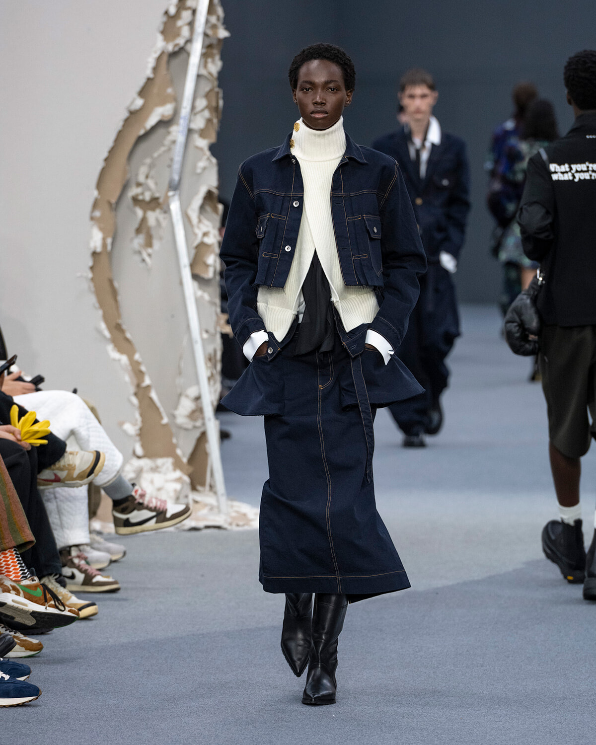 Model walks a fashion show wearing a navy denim jacket over a white turtleneck and a matching denim skirt with black knee-high boots under bright runway lights.