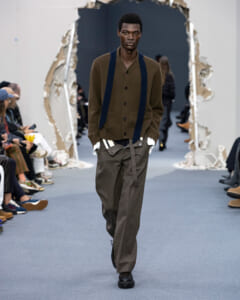 Male model walking a runway in a brown cardigan with navy suspenders and loose olive-gray trousers, audience on both sides