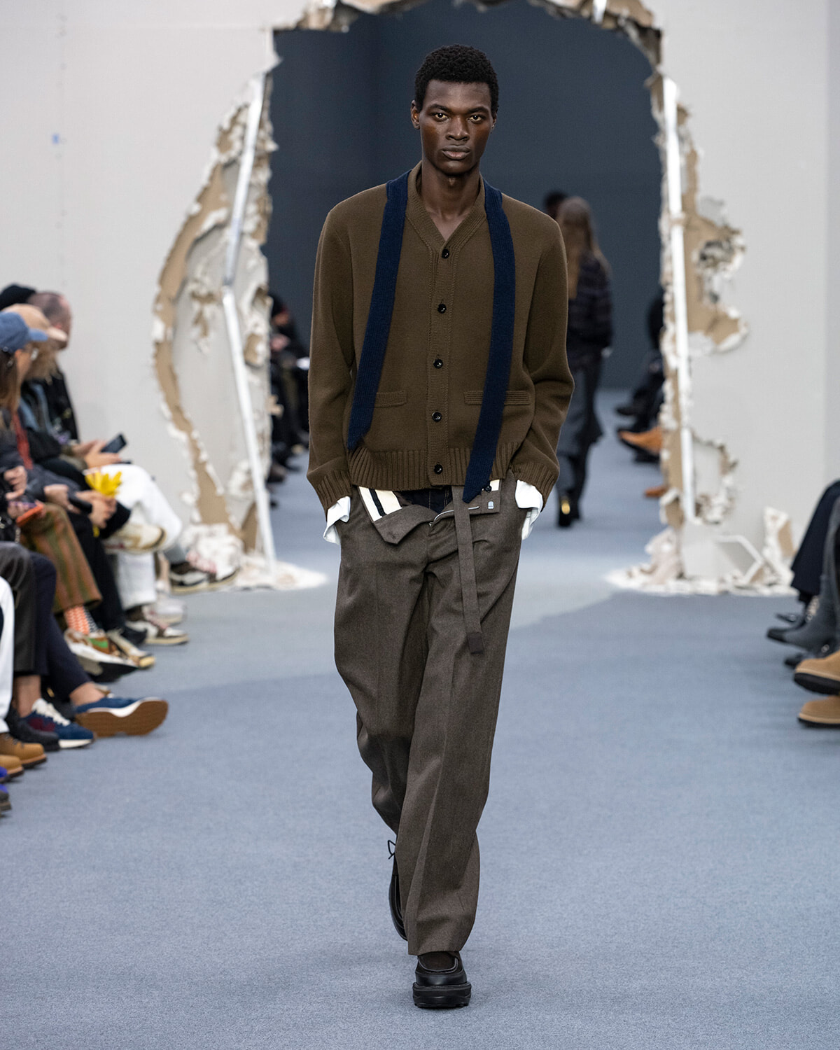 Male model walking a runway in a brown cardigan with navy suspenders and loose olive-gray trousers, audience on both sides