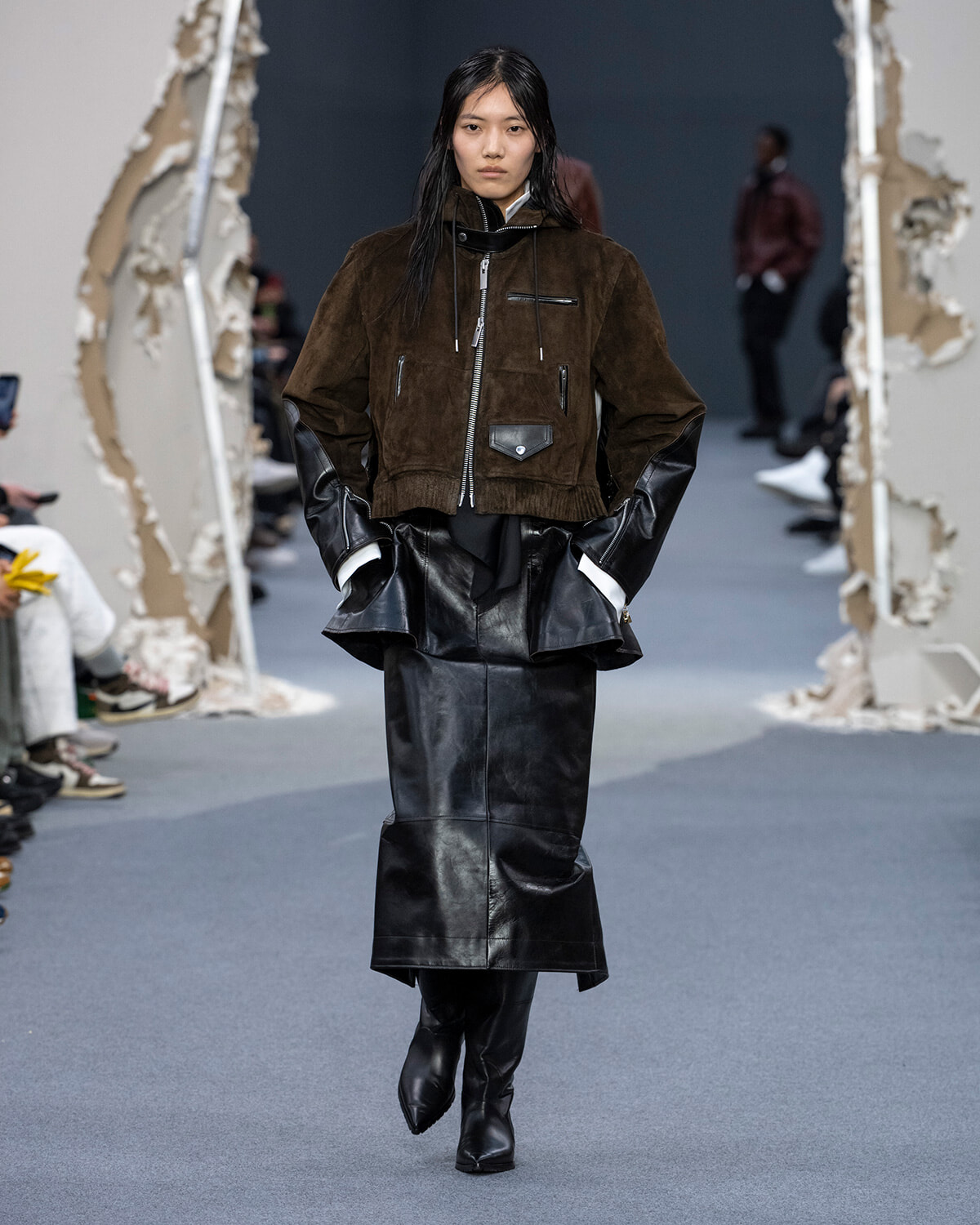 Model walking a fashion runway in a brown suede cropped jacket with black leather skirt and knee-high boots, hands in pockets.